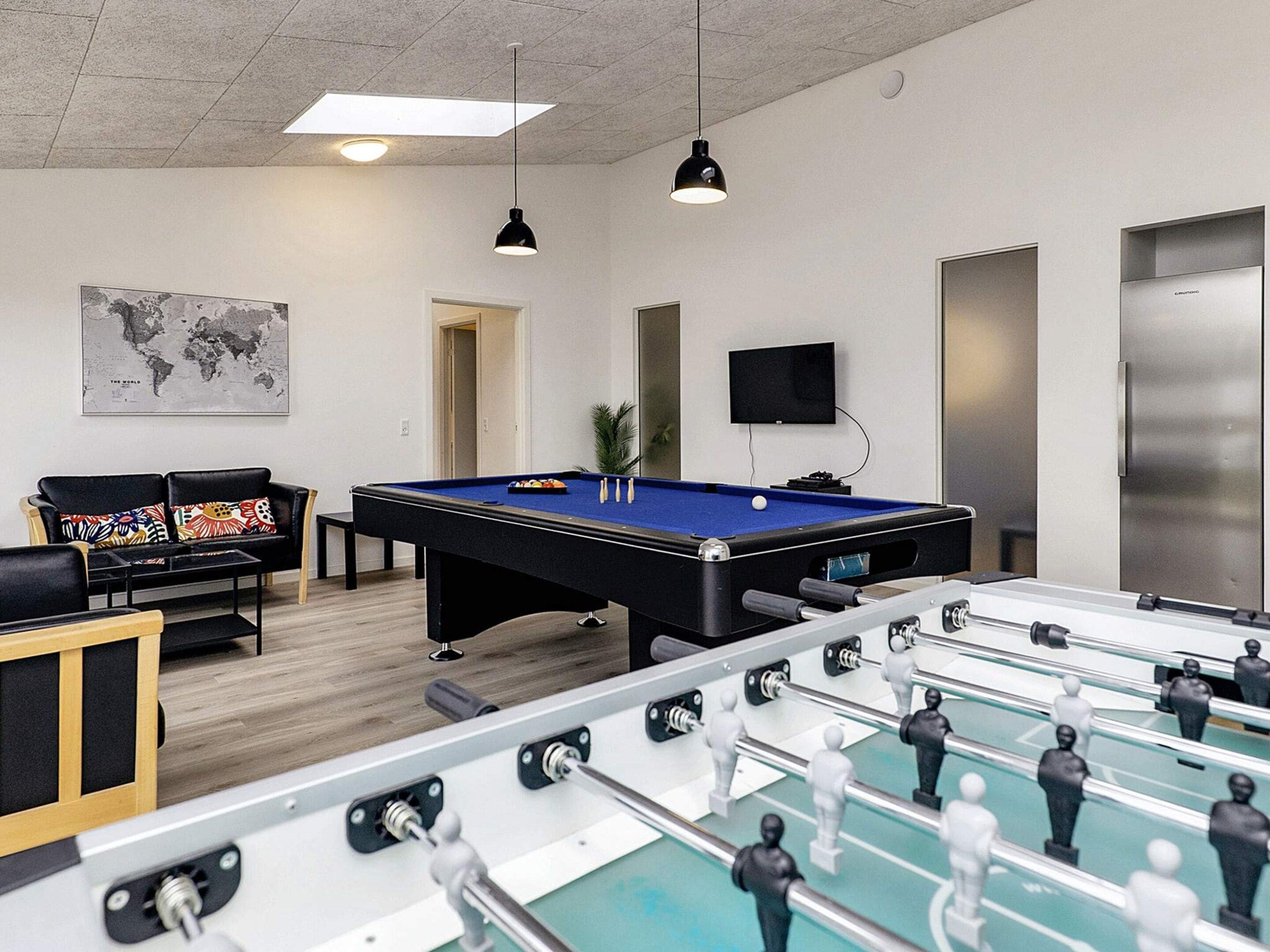 A pool table with a foosball table in the foreground, a sofa with colorful cushions, a world map on the wall, and a television mounted above the pool table.