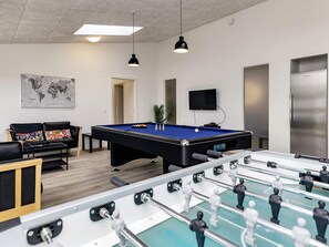 A pool table with a foosball table in the foreground, a sofa with colorful cushions, a world map on the wall, and a television mounted above the pool table.