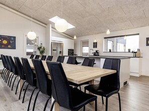 A modern dining area with a long table and chairs, a kitchen in the background, and pendant lighting.