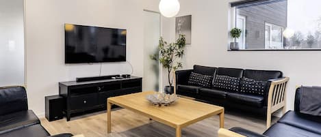 A modern living room with a flat-screen TV, a wooden coffee table, black sofas, and a large window.