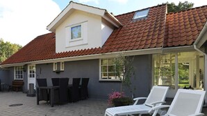 Une maison avec un toit en tuiles, une terrasse avec une table et des chaises, et deux fauteuils de jardin.