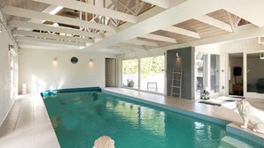 Une piscine intérieure avec un sol carrelé, un plafond en bois et une statue blanche placée sur le bord.