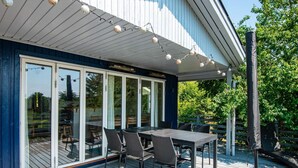A covered outdoor dining area with a table and chairs, a string of lights, and a view of the surrounding greenery.