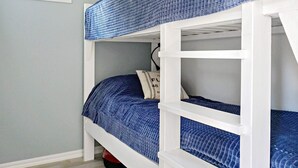 Un lit superposé avec un matelas bleu et une structure blanche, un oreiller orné d’un motif, et une fenêtre donnant sur un paysage.