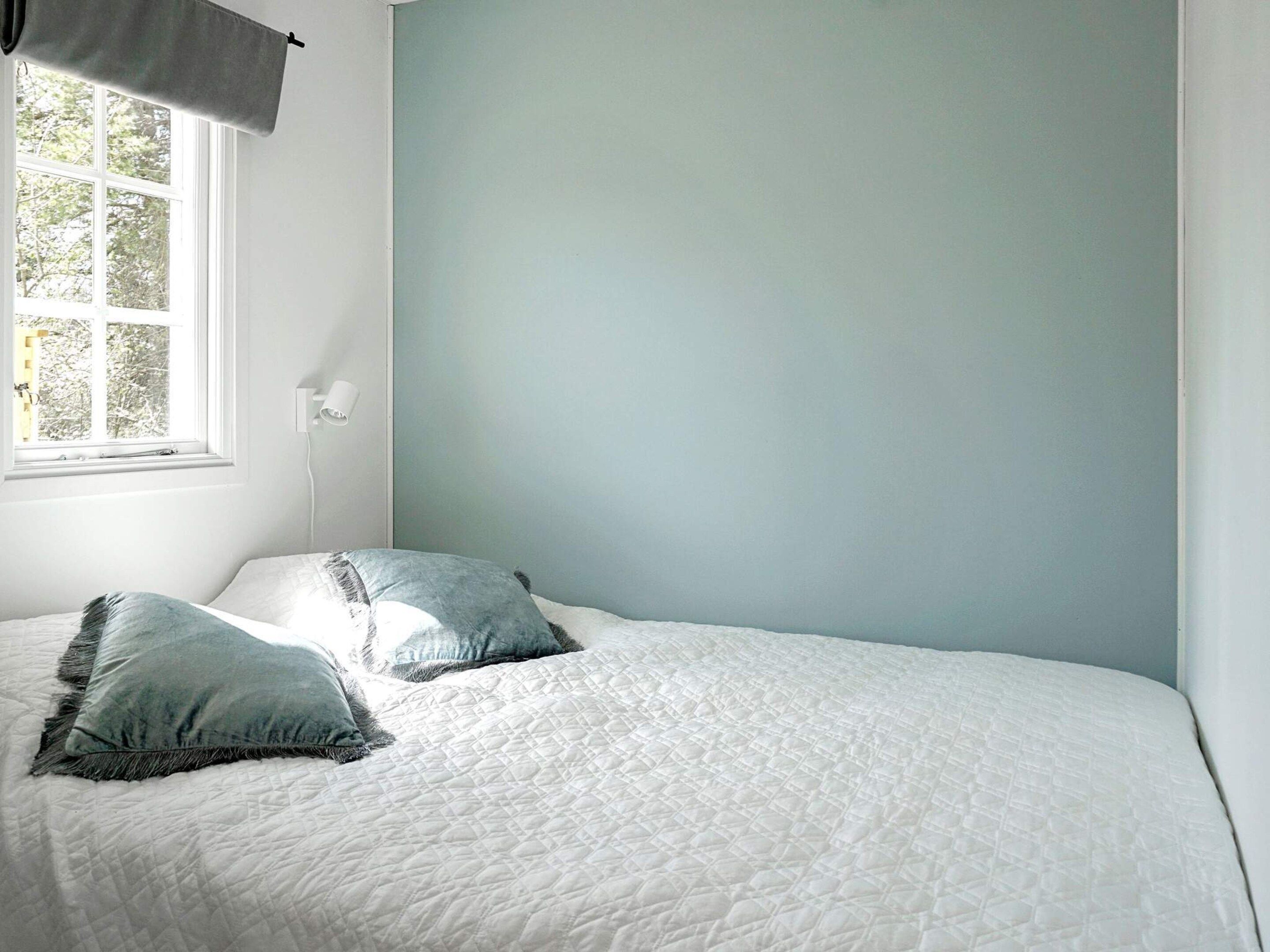 A bedroom with a bed, two pillows, a window with a curtain, and a wall-mounted light.
