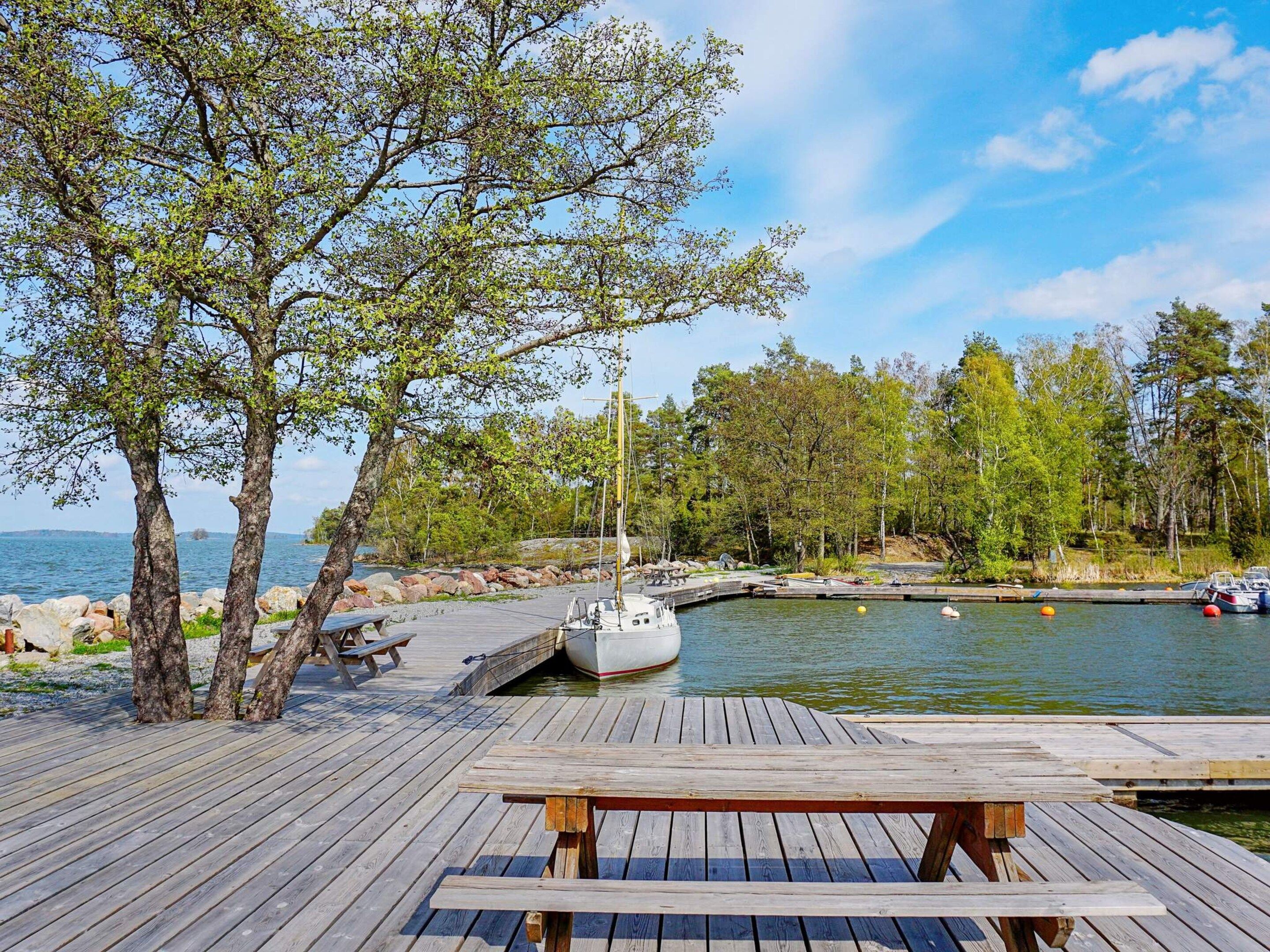 A wooden dock with a picnic table, a sailboat docked at a marina, and a calm lake with trees in the background.