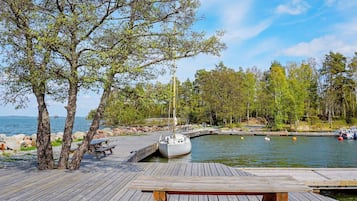 Un muelle de madera con una mesa de picnic, un velero amarrado en un puerto deportivo y un lago tranquilo con árboles al fondo.
