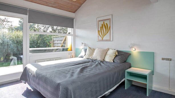 A modern bedroom with a large bed, a bedside table, a nightstand, a window with a view of greenery, and a wall-mounted light fixture. - Holiday Home in VÃ¦ggerlÃ¸se (Vaeggerlose)