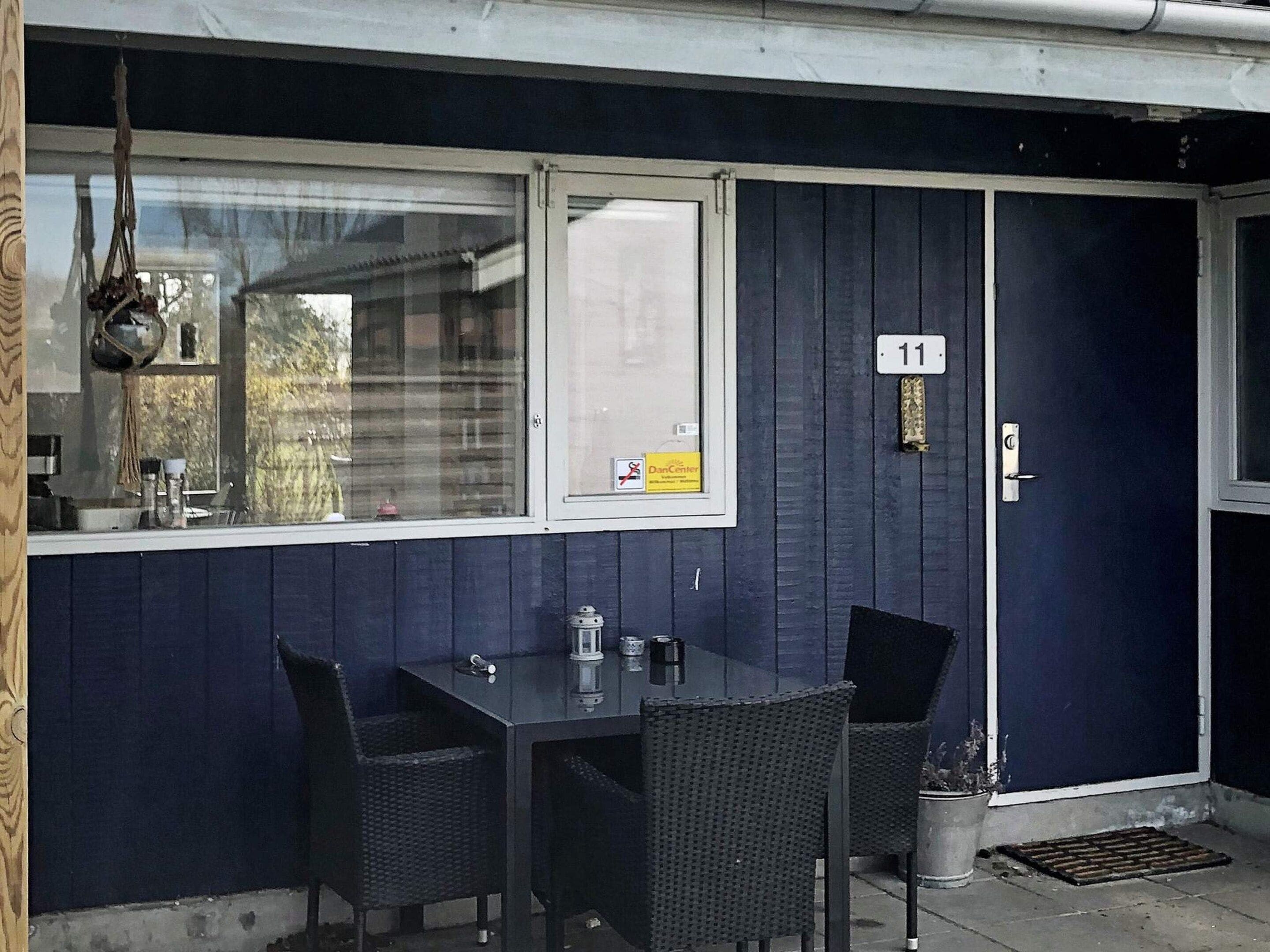 A dark blue exterior wall with a white-framed window, a door with a number plate, and a small table with two chairs.