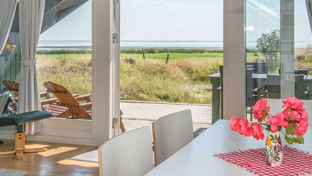 Une pièce dotée d’une grande porte-fenêtre coulissante qui donne sur un espace extérieur offrant une vue sur la mer. On y trouve une table avec un vase de fleurs et une nappe à carreaux.