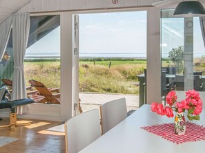 A room with a large sliding glass door leading to an outdoor area with a view of the sea. There is a table with a vase of flowers and a checkered tablecloth. - 6 Person Holiday Home in Romo (Rømø)