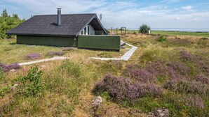 Un bâtiment moderne, de plain-pied, doté d’un toit en pente et d’une cheminée, entouré d’un champ verdoyant parsemé de bruyère violette.