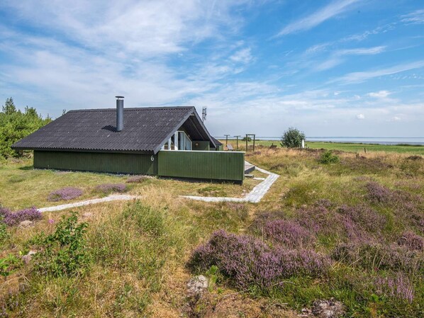 A modern, single-story building with a sloped roof and a chimney, surrounded by a grassy field with purple heather. - 6 Person Holiday Home in Romo (Rømø)