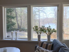 A room with a window overlooking a forest and a body of water, a white table, and a gray sofa with patterned cushions. - 4 Person Holiday Home in Skanes-fagerhult (Skånes Fagerhult)