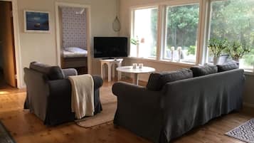 A cozy living room with a sofa, a TV, a small table, and a window with a view of greenery.