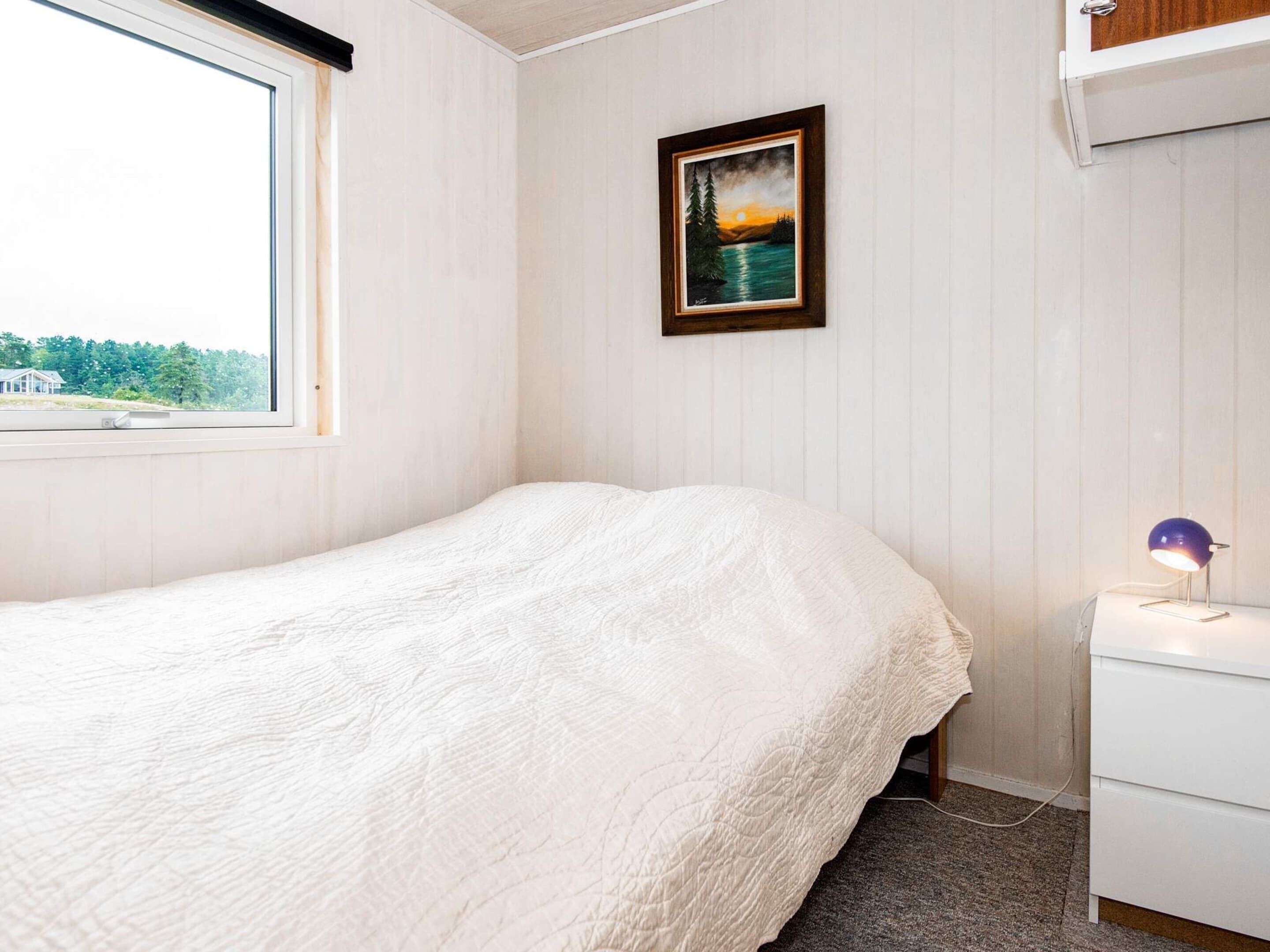 Een slaapkamer met een bed, nachtkastje, lamp en een schilderij aan de muur.