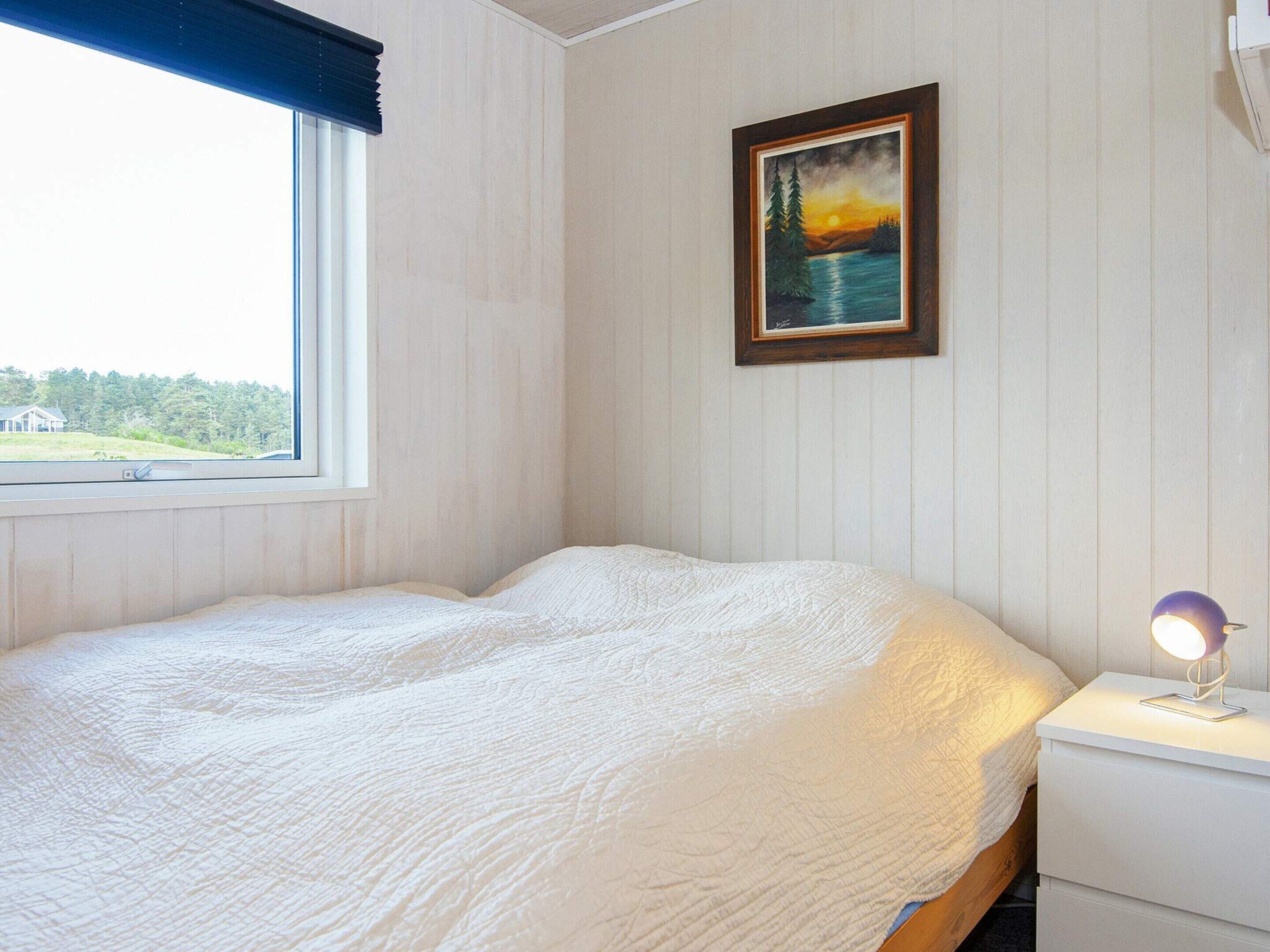 A bedroom with a bed, a nightstand, a lamp, a window with a view of greenery, and a painting on the wall.