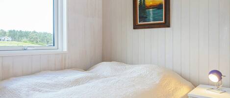 A bedroom with a bed, a nightstand, a lamp, a window with a view of greenery, and a painting on the wall.