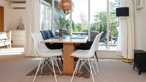 A dining area with a wooden table, white chairs, a hanging light fixture, and large windows with curtains. - Holiday Home in Ebeltoft (Ebeltoft)