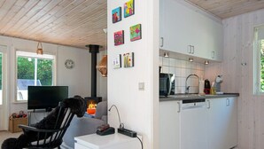 A modern kitchen with white cabinets, a built-in microwave, and a sink. There's a black rocking chair with a fluffy toy in the living area, a flat-screen TV, and a fireplace. - Holiday Home in Ebeltoft (Ebeltoft)