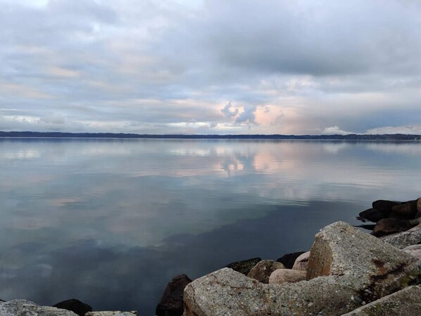 Un lac paisible, bordé d’une rive rocheuse, avec une chaîne de montagnes lointaine se découpant sous un ciel nuageux.