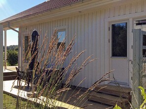 A white building with a tiled roof, a front door, and a window. There is a wooden deck in front of the building. - Holiday Home in Brastad (Brastad)
