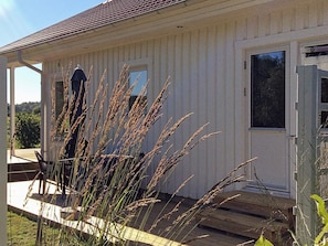 A white building with a tiled roof, a front door, and a window. There is a wooden deck in front of the building. - Holiday Home in Brastad (Brastad)