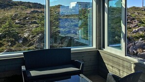 A room with a large window offering a view of rocky terrain and a distant building. There is a small table and two chairs inside the room. - Holiday Home in Dyrvik (Frøya)