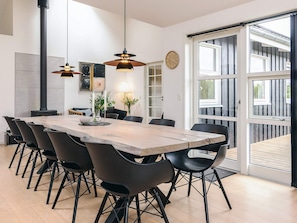 A modern dining area with a long wooden table, black chairs, pendant lights, and a large window with blinds.