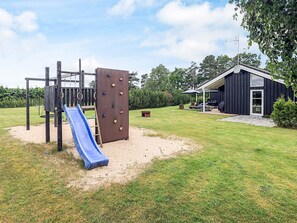 Un parque infantil con tobogán y pared de escalar, una zona de barbacoa y un edificio con porche cubierto.