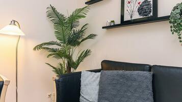 A modern living room with a black leather sofa, a large green plant, a floor lamp, and a framed artwork on the wall.