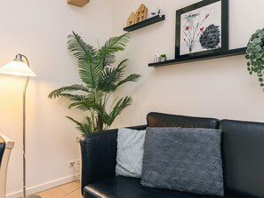 A modern living room with a black leather sofa, a large green plant, a floor lamp, and a framed artwork on the wall.