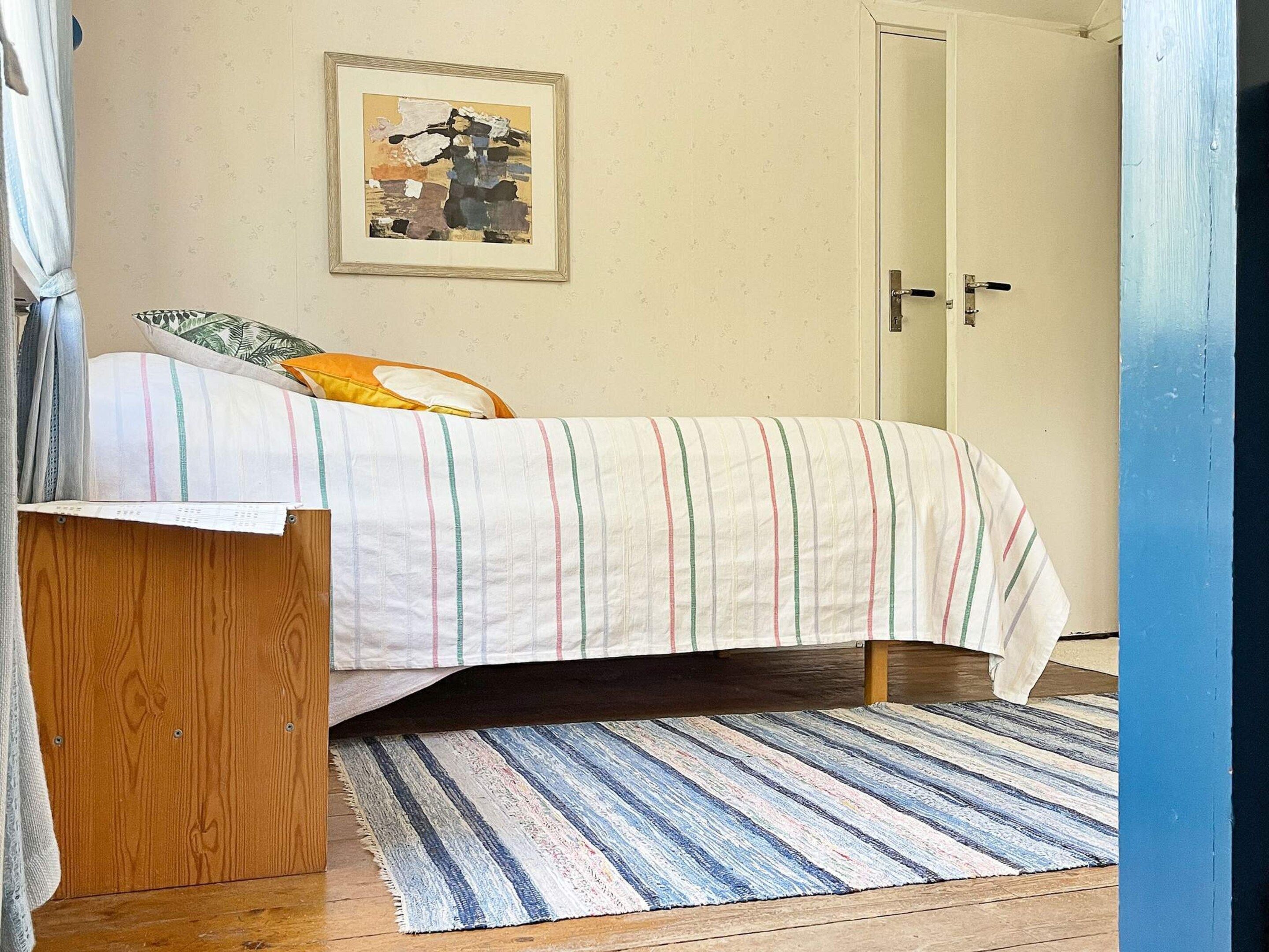 A bedroom with a bed, a wooden nightstand, a striped rug, a door, and a framed picture on the wall.