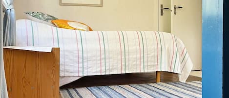 Un dormitorio con cama, una mesita de noche de madera, un tapete a rayas, una puerta y un cuadro en la pared.