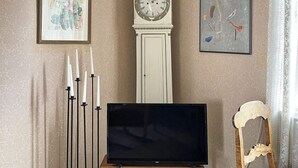A room with a flat-screen TV on a wooden stand, a tall white clock, and two framed artworks on the walls. - 5 Person Holiday Home in Linneryd/kronobergs Lan (Linneryd)