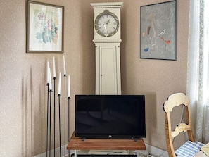 Een kamer met een flatscreen televisie op een houten standaard, een hoge witte klok en twee kunstwerken in lijsten aan de wanden.