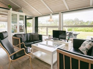 A modern living room with a black leather sofa, a white coffee table, and a television. There is a glass cabinet with decorative items and a view of the outdoors through large windows.