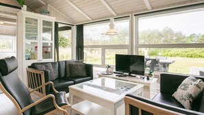 A modern living room with a black leather sofa, a white coffee table, and a television. There is a glass cabinet with decorative items and a view of the outdoors through large windows. - Holiday Home in Løkken (Lokken)
