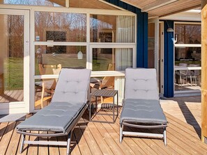 A covered outdoor seating area with two lounge chairs and a table, large windows, and a view of the outdoors. - Holiday Home in Gilleleje (Gilleleje)