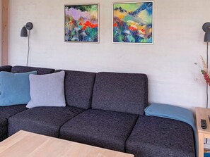 A modern living room with a dark grey sectional sofa, two abstract paintings, a black floor lamp, and a small wooden side table with a remote control. - Holiday Home in Gilleleje (Gilleleje)