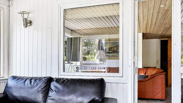 Un canapé en cuir noir se trouve dans une pièce aux murs blancs et au plafond en bois. Une grande fenêtre offre une vue sur un jardin et un bâtiment à l’extérieur.