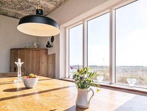 Et spiseområde med et træbord, en hvid kande med blomster og udsigt gennem store vinduer.