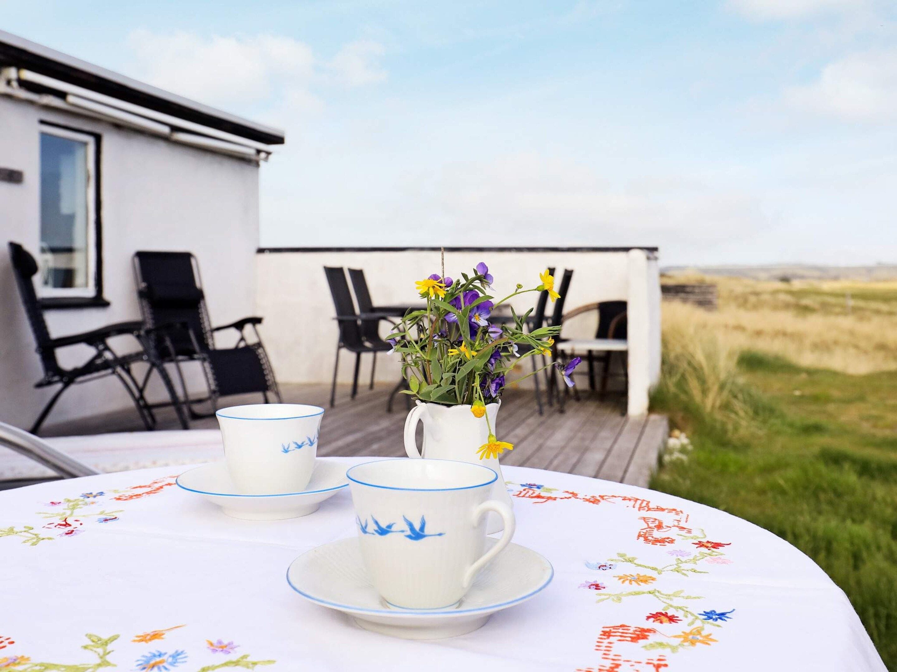 Une table dressée avec des tasses et des soucoupes blanches, un vase rempli de fleurs et une vue sur un bâtiment blanc et un paysage verdoyant.