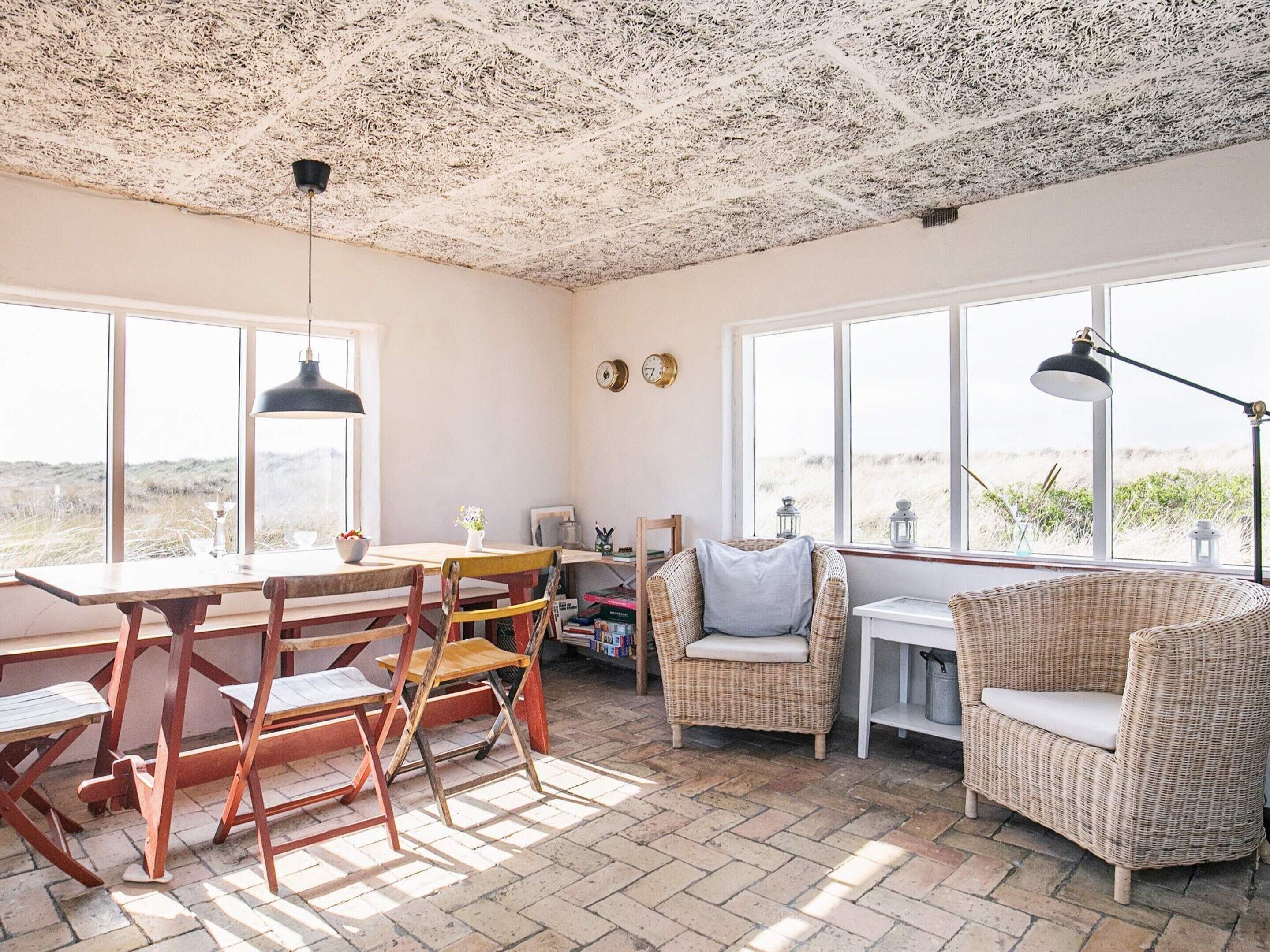 A cozy room with a rustic ceiling, large windows offering a view of the beach, a dining table with chairs, and a wicker sofa.