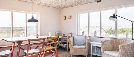 Ett mysigt rum med ett rustikt tak, stora fönster med utsikt över stranden, ett matbord med stolar och en soffa av rotting.