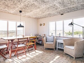 A cozy room with a rustic ceiling, large windows offering a view of the beach, a dining table with chairs, and a wicker sofa.