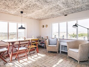Une pièce chaleureuse avec un plafond rustique, de grandes fenêtres donnant sur la plage, une table à manger avec des chaises et un canapé en osier.