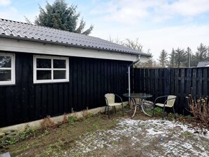 Et sort hus med tagsten, to vinduer, og en lukket baghave med et bord og stole.