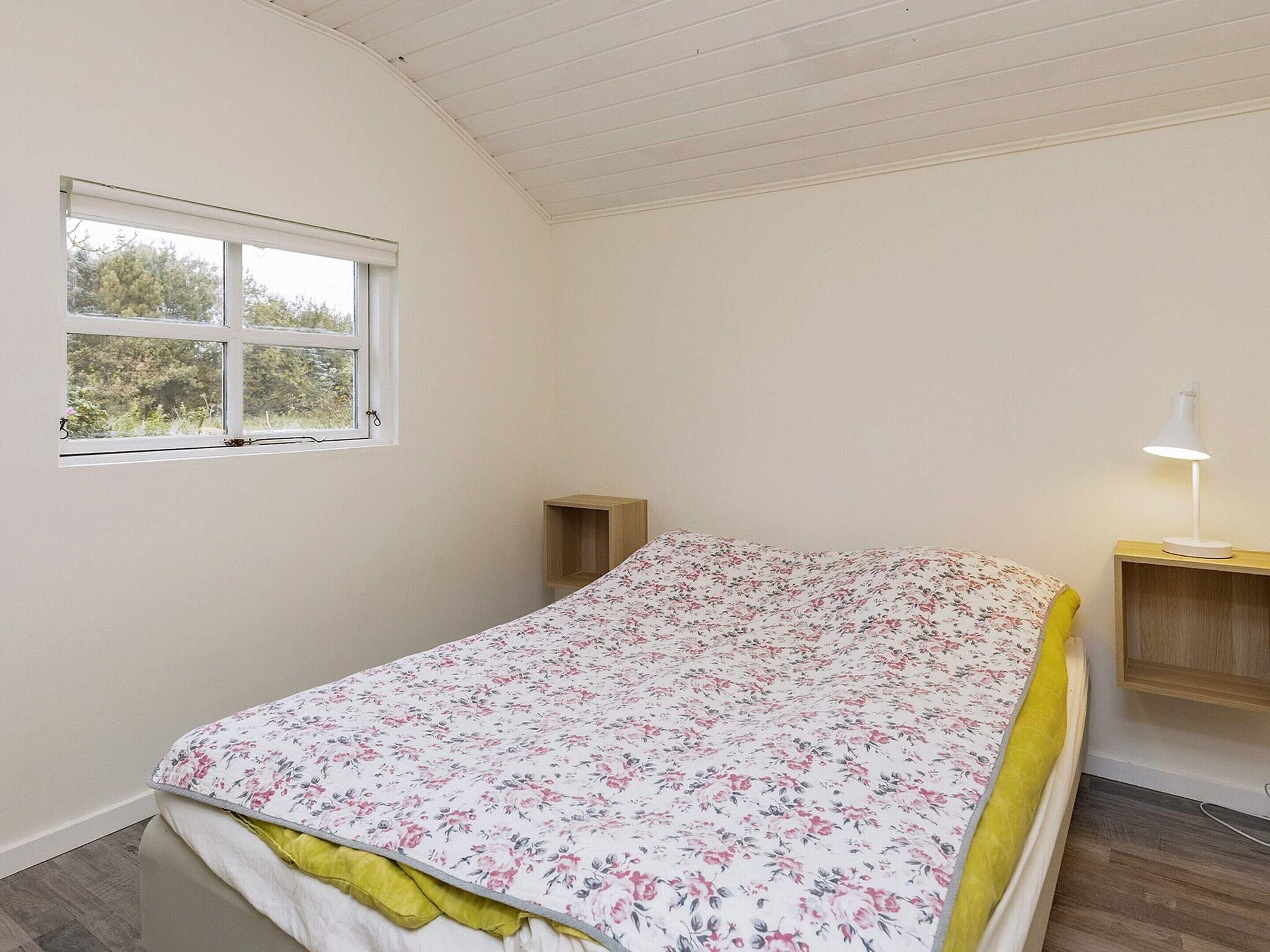 A bedroom with a bed, a window, a bedside table with a lamp, and a wooden nightstand.