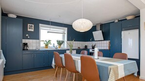 A compact kitchen with dark blue cabinets, a wooden dining table with striped tablecloth, and a hanging light fixture. - 7 Person Holiday Home in Lemvig (Lemvig)